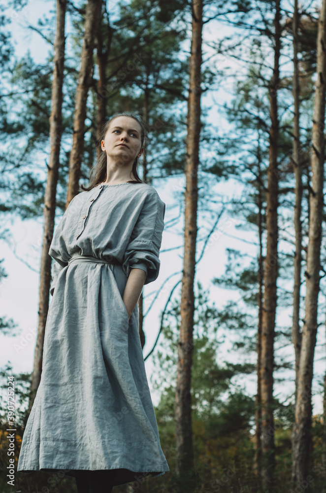 Side view moody portrait of a young, thoughtful, meditative, caucasian woman in a long, grey, organic, linen dress leaning to the side enjoying the forest bath in a pine tree forest Side view moody portrait of a young, thoughtful, meditative, caucasian woman in a long, grey, organic, linen dress leaning to the side enjoying the forest bath in a pine tree forest