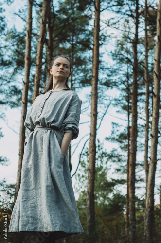 Side view moody portrait of a young, thoughtful, meditative, caucasian woman in a long, grey, organic, linen dress leaning to the side enjoying the forest bath in a pine tree forest