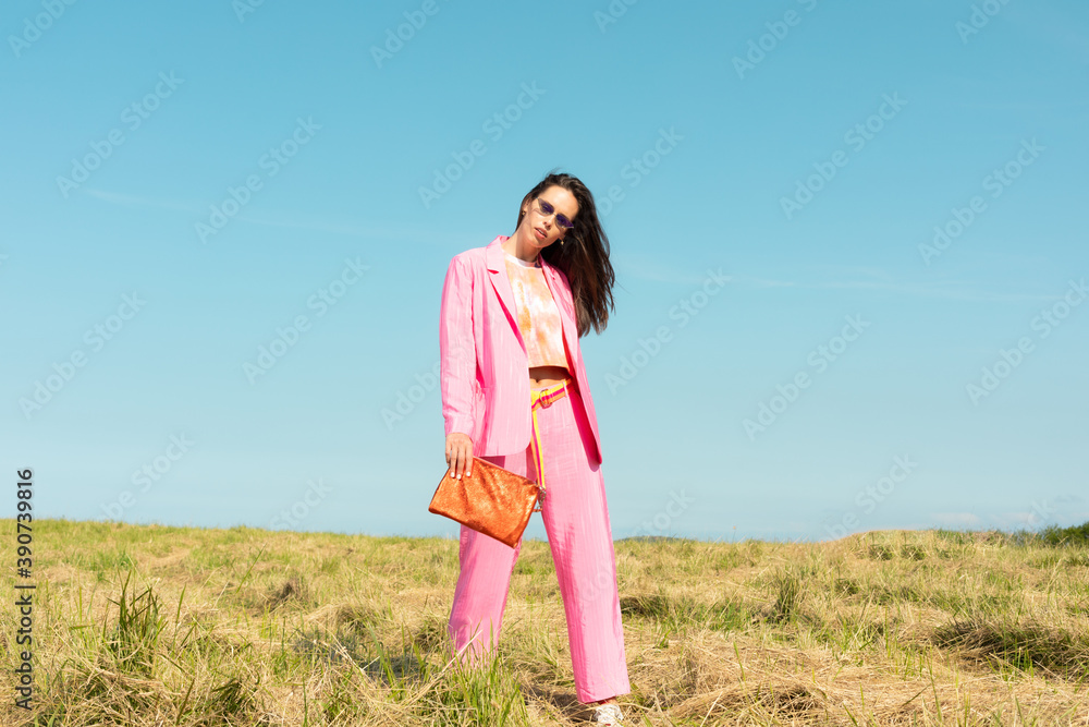 
Young model woman wearing fashionable pink pants and glasses posing by the grass and blue sky playing with her long hair. Cute modern European model girl enjoys a walk on the beach. Spring summer 