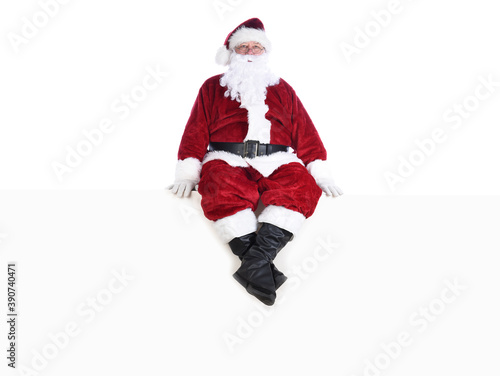 Senior man in traditional Santa Claus Suit sitting on a white wall.  Isolated on white with copy space.