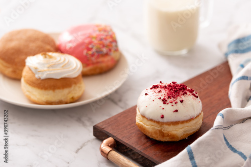 Close up of a donut with white icing and red decorations on a board with a group of mixed donuts arranged on a plate behind and a glass of milk. Ideal for recipe page or magazine editorial