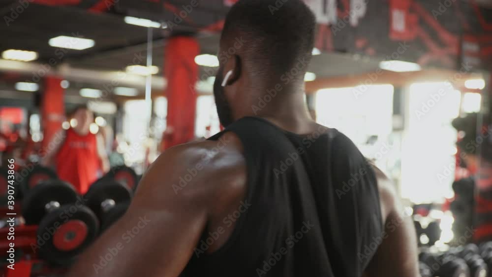 Back view of african american man bodybuilder with wireless headphones performing Side raise exercise with dumbbells at gym Back view of african american man bodybuilder with wireless headphones performing Side raise exercise with dumbbells at gym