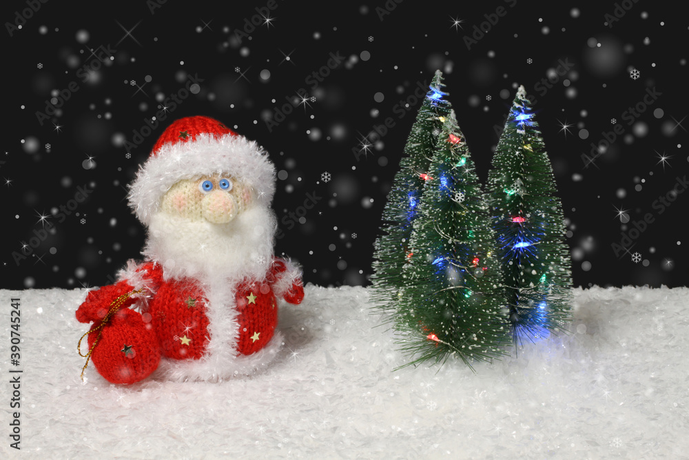The toy knitted wool Santa Claus stands next to the toy green Christmas trees with lights on artificial white snow against black background with snow mist. Closeup