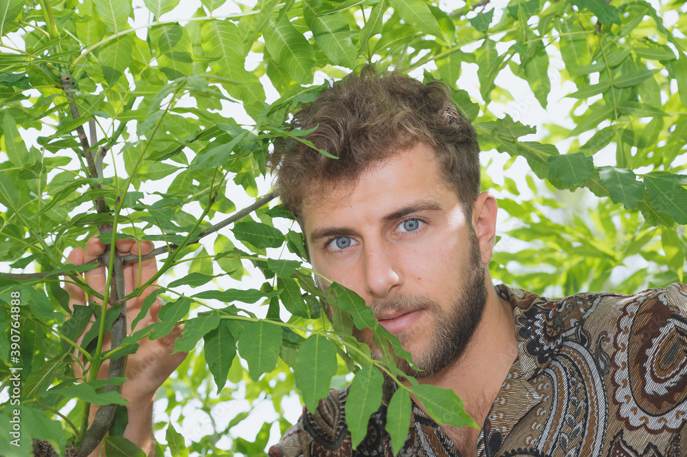 Close up portrait of young millennial man with hazel curly hair and beard wearing casual dark motley shirt. Natural neutral skin tone. Walks around the park hiding behind the leaves, bright sunny day Close up portrait of young millennial man with hazel curly hair and beard wearing casual dark motley shirt. Natural neutral skin tone. Walks around the park hiding behind the leaves, bright sunny day