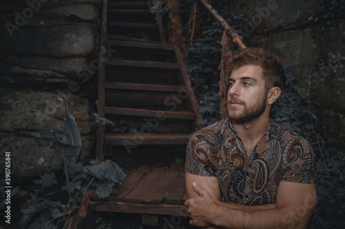 Portrait of strong muscle fit young curly man with brown hazel hair and beard wearing casual motley shirt. In the forest near wooden stairs. Dark natural skin tone, moody. Cheerful male adult journey.