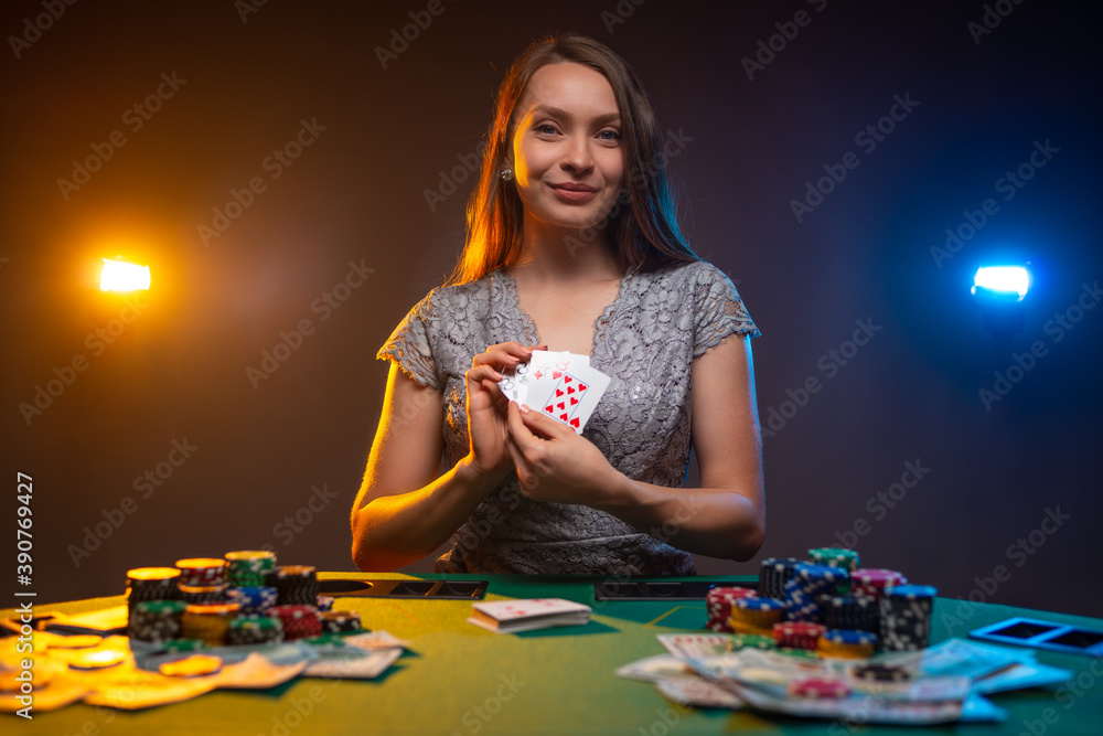 Gambler happy woman shows her cards at poker table in casino on dark smoky background with lamps light. Emotional face. Gambling concept. Gambler happy woman shows her cards at poker table in casino on dark smoky background with lamps light. Emotional face. Gambling concept.