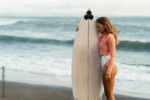 Young attractive surfer woman with white board at sunset on the ocean. Bali Indonesia. Summer time, sports, travel content.