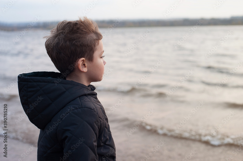 a blond boy in a dark jacket stands in the wind on the Bank of the river and looks into the distance