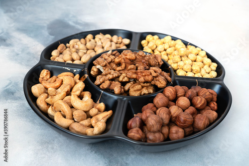 A large serving platter with five compartments in assorted shelled nuts (hazelnuts, cashews, peanuts, walnuts) and chickpeas stands on a light gray concrete background. Healthy food, vitamins