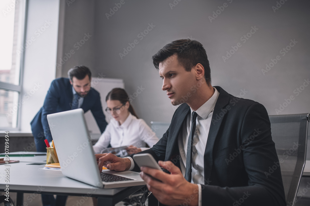 Man with smartphone looking at laptop, colleagues behind Man with smartphone looking at laptop, colleagues behind