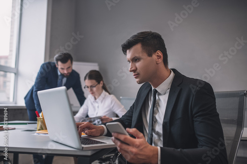 Man with smartphone looking at laptop, colleagues behind