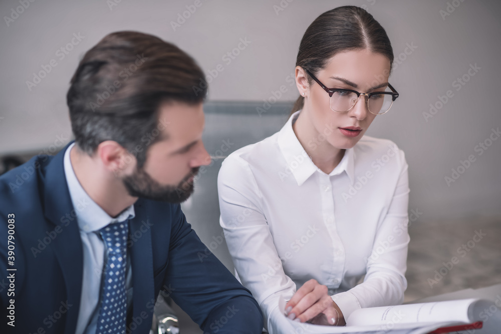 Bearded male and brown-haired female in eyeglasses sitting at table, reading papers Bearded male and brown-haired female in eyeglasses sitting at table, reading papers