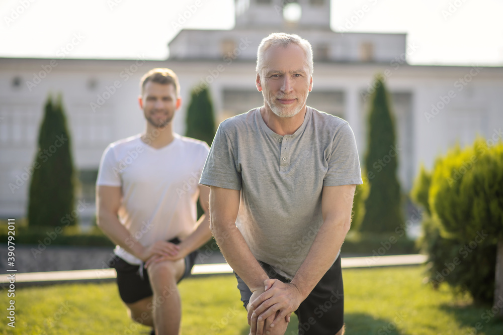 Brown-haired male and grey-haired male stretching legs outside, hands on knee Brown-haired male and grey-haired male stretching legs outside, hands on knee