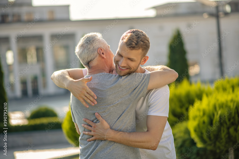 Brown-haired male and mature grey-haired male embracing each other, smiling Brown-haired male and mature grey-haired male embracing each other, smiling