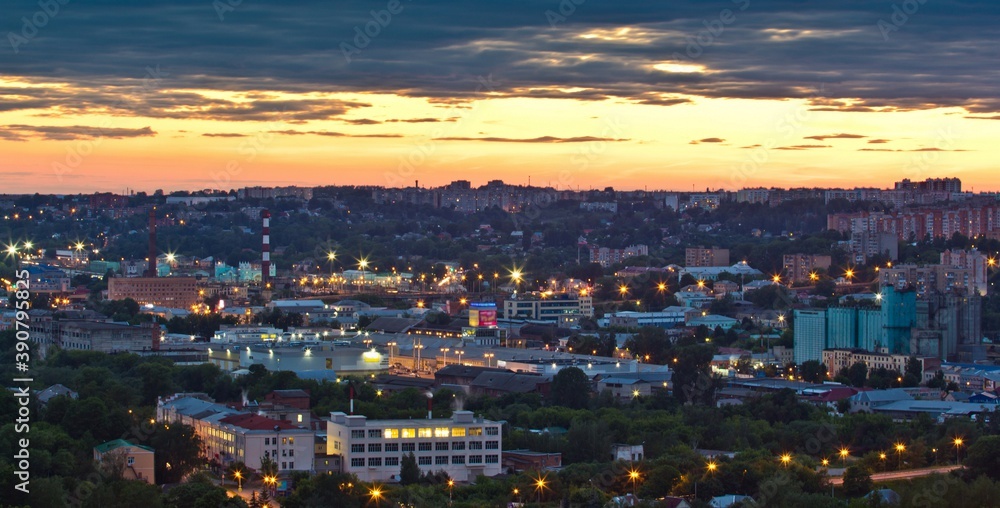 Cityscape of Smolensk at the sunset: lit up street lamps among buildings and houses and cloudy orange sky over it in Russia.