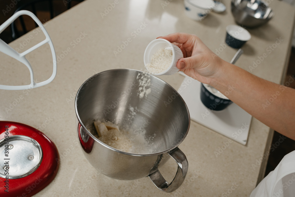 A close photo of the hand of a young woman who is putting an almond flour from the cup into the stainless steel bowl of the stand mixer. A close photo of the hand of a young woman who is putting an almond flour from the cup into the stainless steel bowl of the stand mixer.