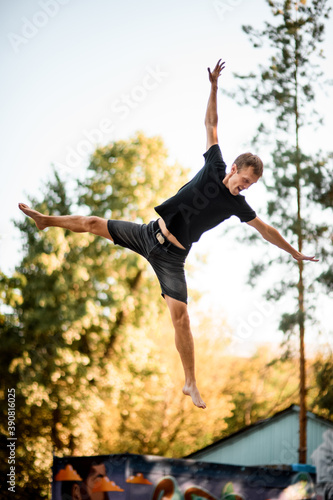 Athletic man skillfully jumps on trampoline against the backdrop of green trees and sky.