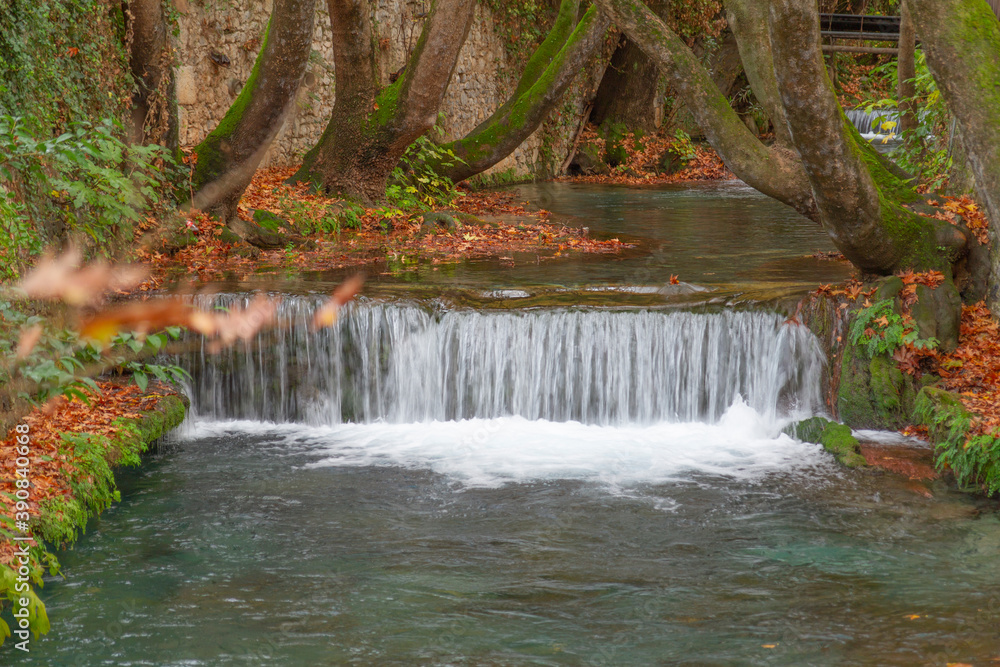 Obraz premium Waterfall in Krya, Livadia Greece. The river flows between the tree trunks. The leaves, green, red and yellow on the ground. The exposure time is 1/25 and the waterflow looks silky 