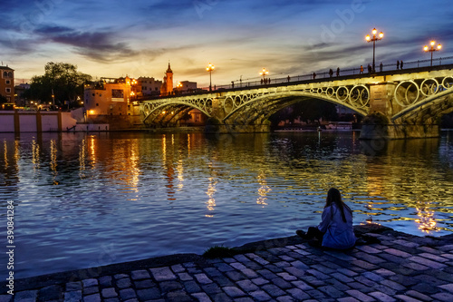 Chica de espaldas sentada en el suelo mirando al río Guadalquivir y al puente de triana al atardecer. photo