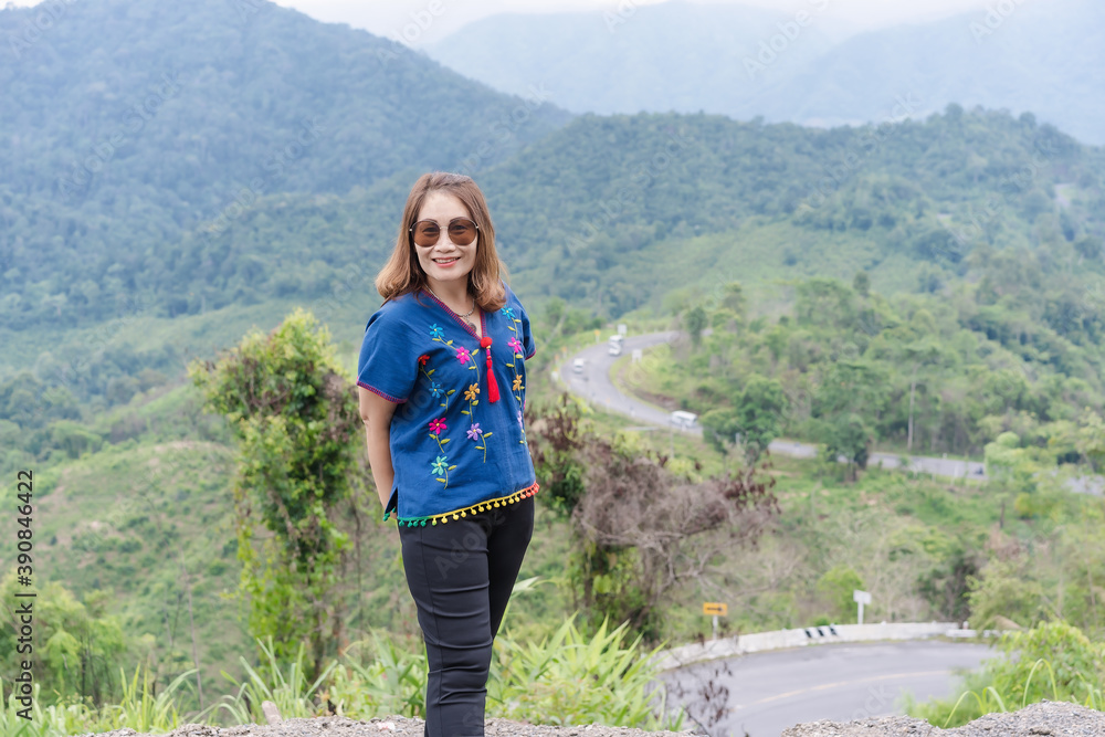 Naklejka premium Adult traveling woman wearing sunglasses standing and smiling on the top of the mountain cliff with relaxing mood and watching beautiful view of woods and clouds foggy on vacation. Nature background