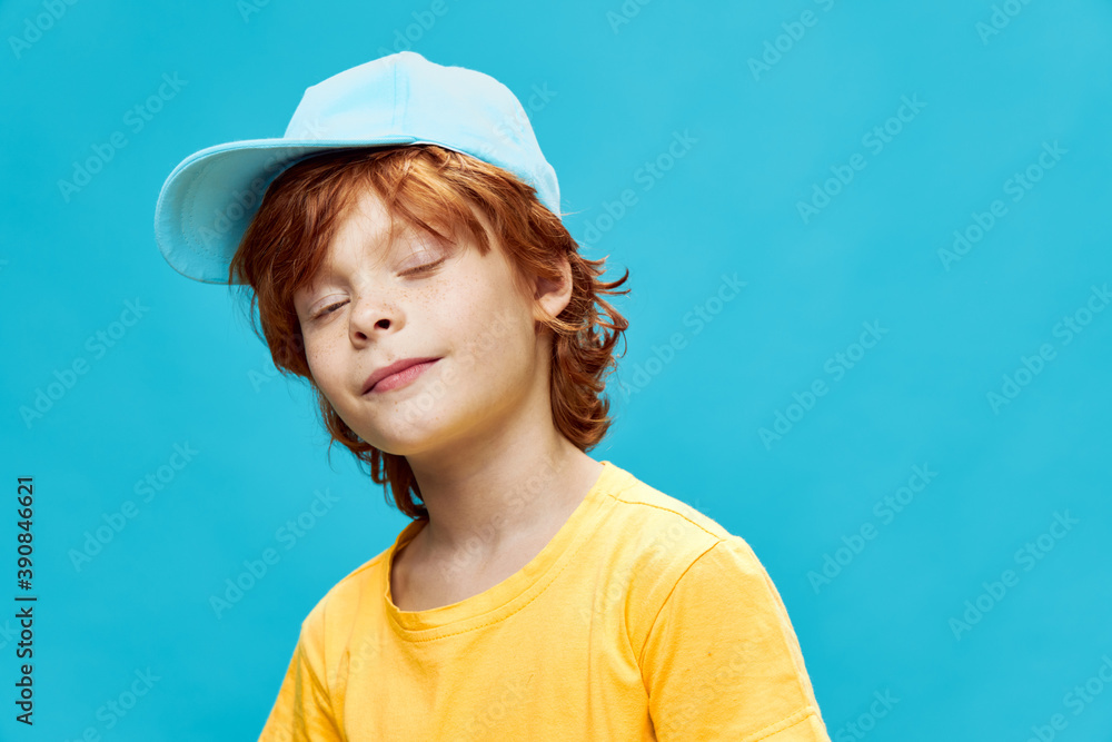 Red-haired boy in a blue cap yellow t-shirt closed eyes blue background Red-haired boy in a blue cap yellow t-shirt closed eyes blue background