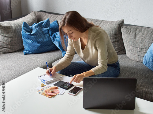 Young woman sitting on the couch in the living room and doing financial balance sheet. The girl counts bills using a calculator. There is euro money and a laptop on the table.  photo