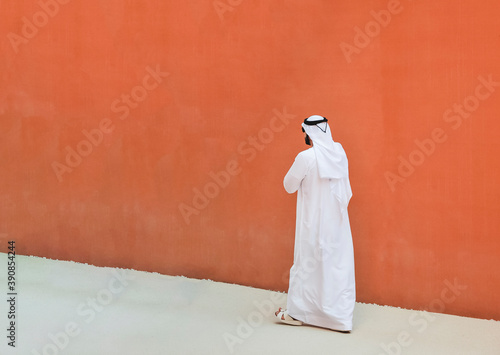 Arabic man with traditional emirates clothes walking outdoors in the street against a orange wall background - Arab ethnicity and muslim religion concept - Copy space for text photo