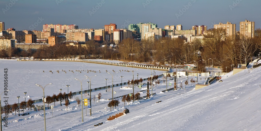 The view on the embankment of frozen Izhevsk pond and on the living block - many tall houses in the background in winter, Russia.
