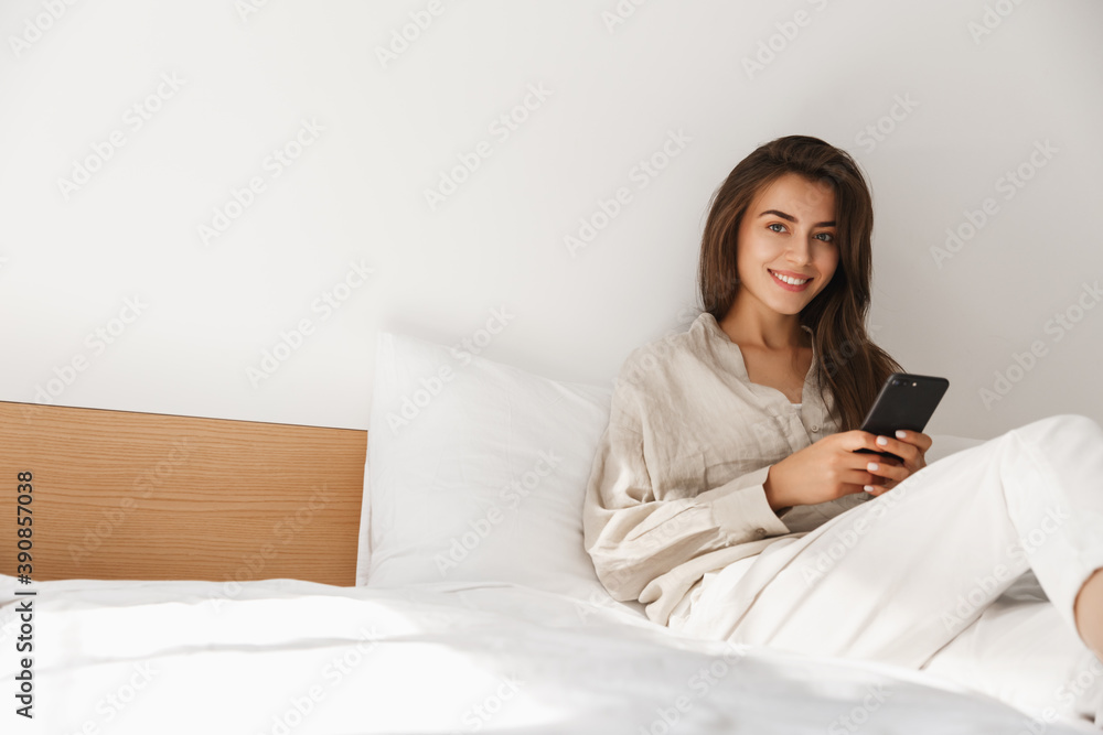 Technology, communication and people concept - happy young woman with smartphone texting message in bed at home. Girl using mobile phone and relaxing in bedroom Technology, communication and people concept - happy young woman with smartphone texting message in bed at home. Girl using mobile phone and relaxing in bedroom