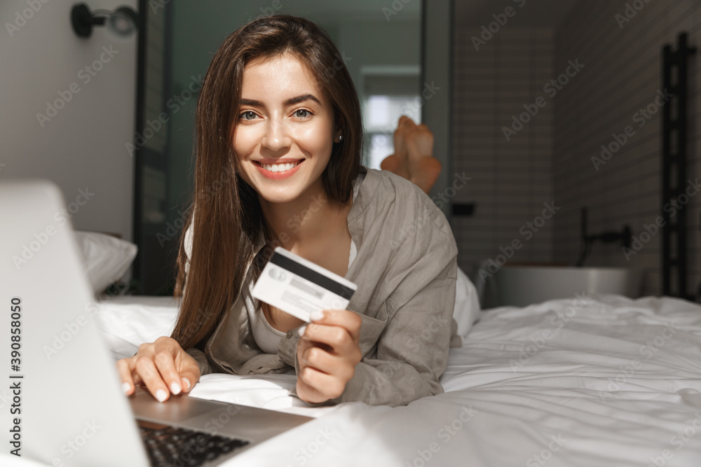 Image of young woman with credit card and laptop shopping in internet, lying in bed and smiling at camera Image of young woman with credit card and laptop shopping in internet, lying in bed and smiling at camera