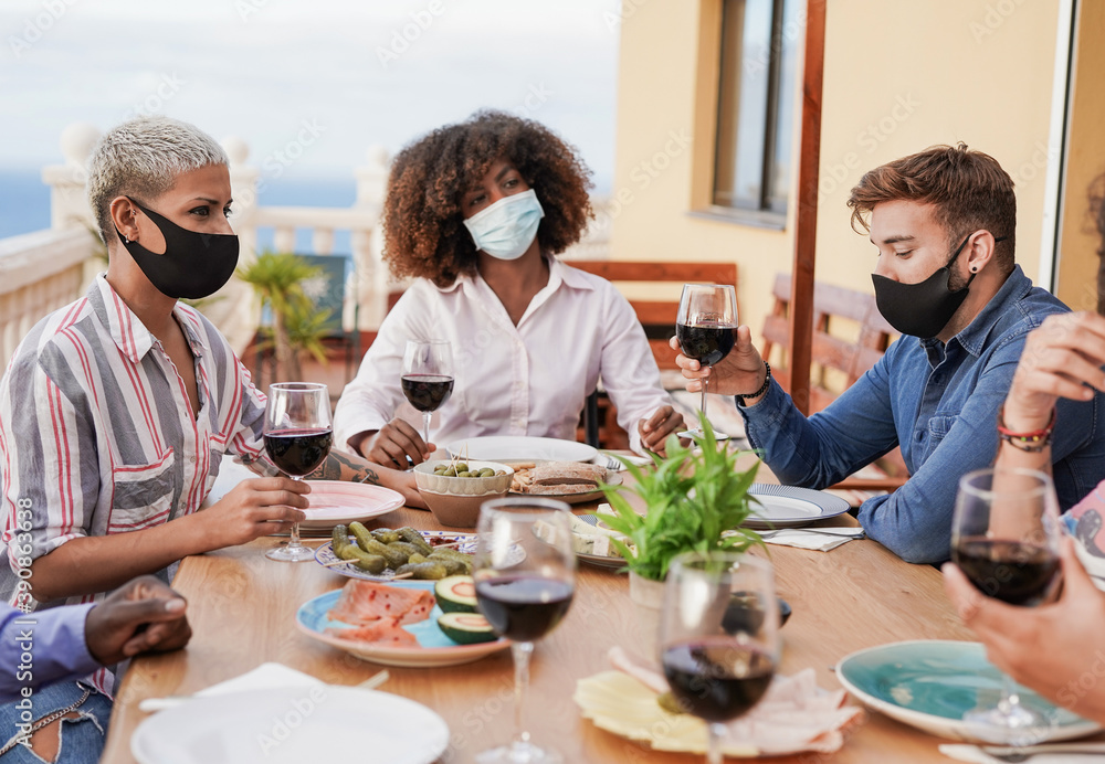 Multiracial people enjoy dinner on patio at home while wearing protective face mask for coronavirus - Prevention and safety measures