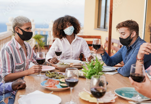 Multiracial people enjoy dinner on patio at home while wearing protective face mask for coronavirus - Prevention and safety measures