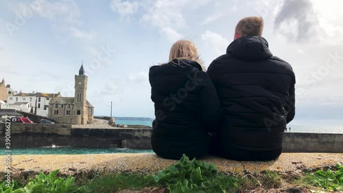 Young Couple Sitting Together And Pointing At Porthleven Clock Tower In Porthleven, Helston, UK. - wide shot photo