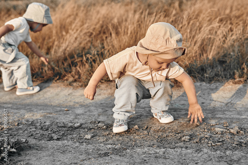 A two-year-old child in a hat crouched touches something on a country road, summer. Experience of the world.
