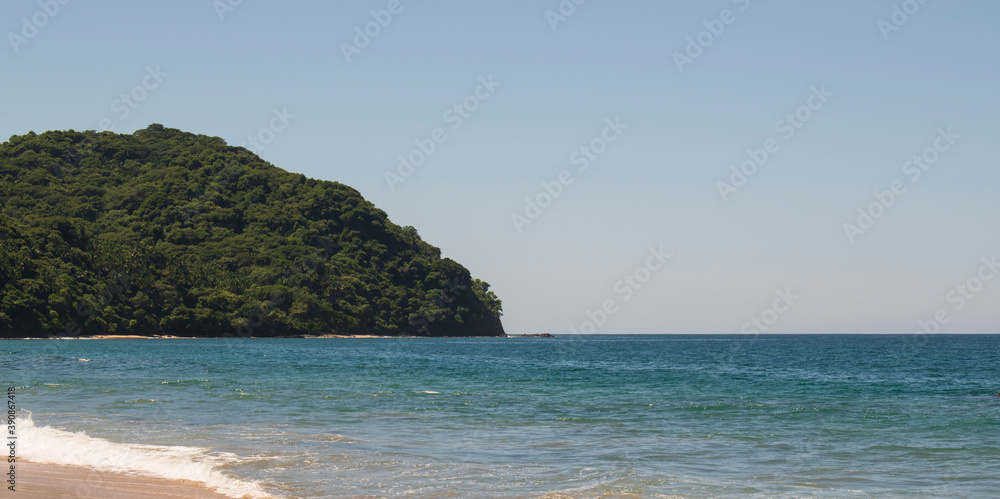 Fototapeta premium Beautiful landscape of a beach in the mexican pacific coast in a sunny day with a blue and clear sky, trees, palms and green hills, some waves and sand in the beach