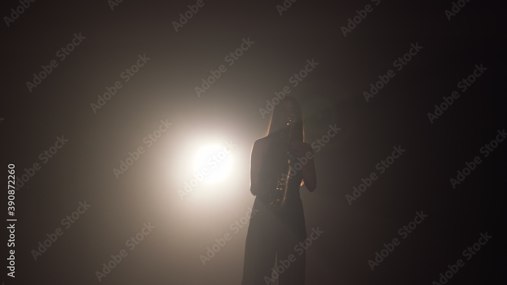 Young beautiful girl in a dark dress plays on a golden shiny saxophone on stage. Dark studio with smoke and stage lighting. Hands and saxophone close up. Side view.Slow motion video Young beautiful girl in a dark dress plays on a golden shiny saxophone on stage. Dark studio with smoke and stage lighting. Hands and saxophone close up. Side view.Slow motion video