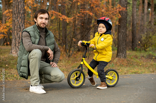 Happy young father squatting by his adorable little son sitting on balance bike