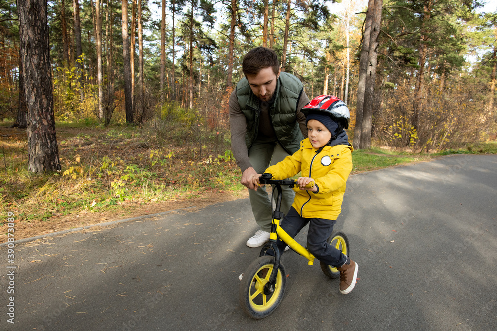Young father slightly bending while helping his little son to ride balance bike Young father slightly bending while helping his little son to ride balance bike