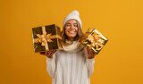 young smiling happy pretty woman holding golden present boxes celebrating new year, christmas gifts, wearing white knitted sweater, scarf and hat, winter fashion trend, posing on yellow background