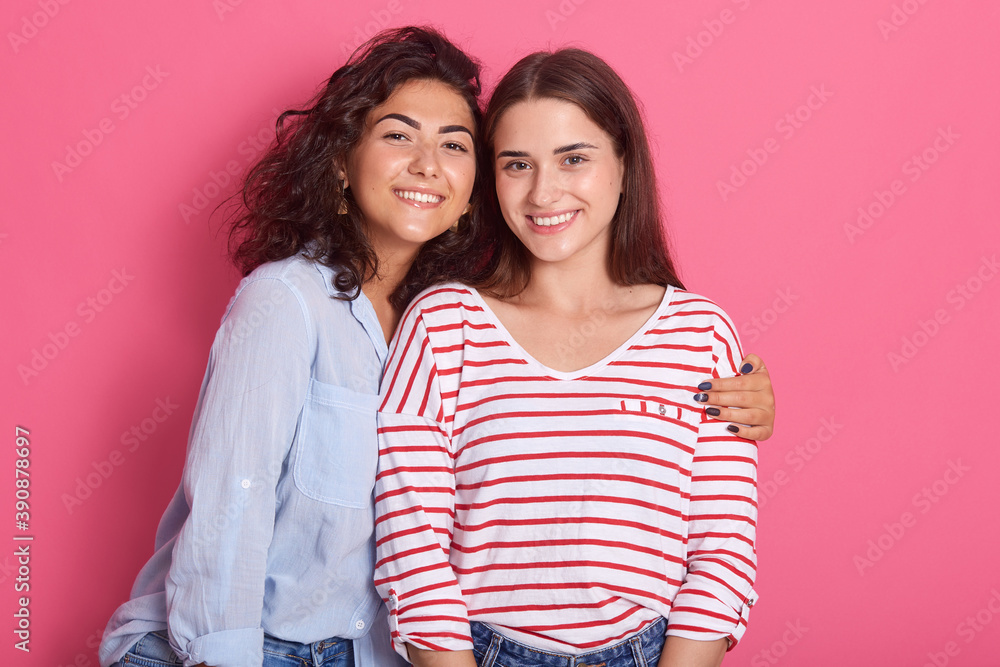 Two smiling ladies posing with charming smiles, have dark hair, wearing casual clothing, looking at camera and showing their teeth, wavy haired girl hugs her friend, females expressing positive. Two smiling ladies posing with charming smiles, have dark hair, wearing casual clothing, looking at camera and showing their teeth, wavy haired girl hugs her friend, females expressing positive.