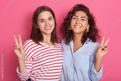 Two girlfriends having fun isolated over pink background, girls look at camera with toothy smile and showing victory sign, expressing happiness while hugging each other, females dress casual attires.