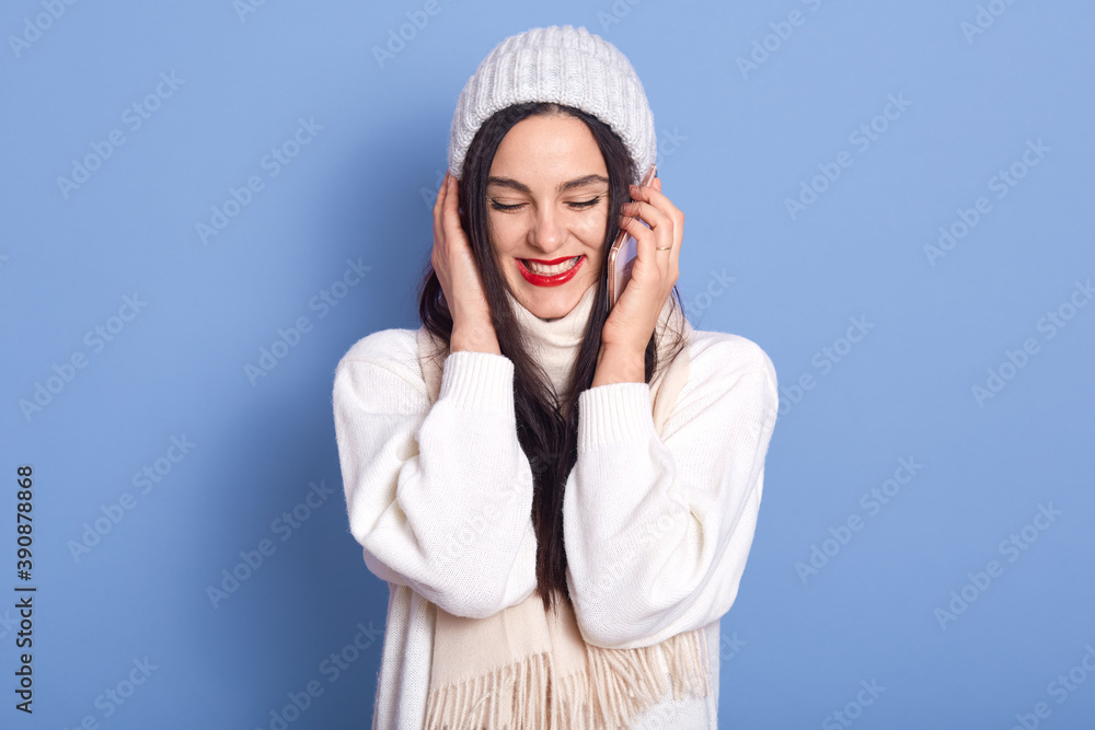 Happy pretty brunette girl talking on phone and laughing, holding smartphone near ear, female dresses white sweater, scarf, cap, standing isolated over blue background, girl wears red lipstick.