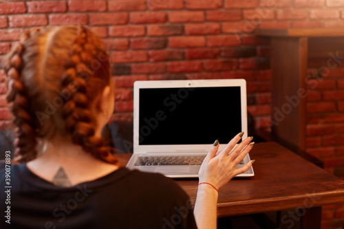 Quadro su tela Young woman sitting at cafe with laptop, showing blank screen of notebook, lady posing backwards against brick wall, girl with pigtails dresses black t shirt, female spreading hand with manicure
