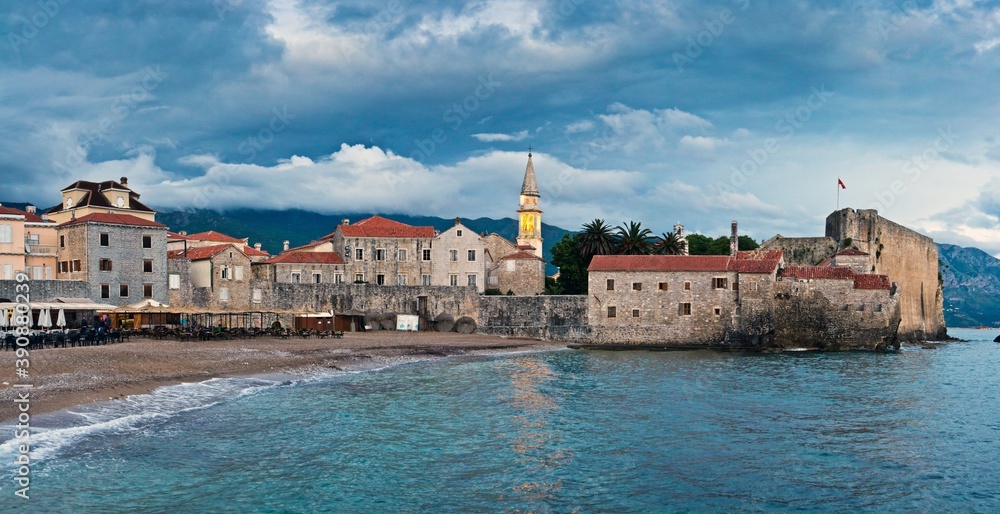 Grey houses with tile roofs and a cafe on the coast of Adriatic Sea in the old town of Budva and mountains in clouds in the background in Montenegro.