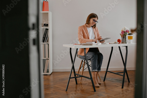 Positive businesswoman in stylish outfit with smile working in tablet, sitting at bright desktop