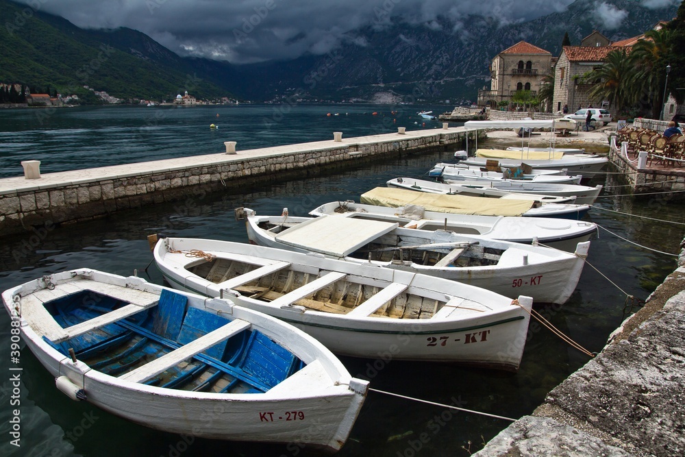 Many white wooden boats moored in a row to a dock in Perast, Montenegro. An embankment with medieval stone houses of the Bay of Kotor and green mountains in the background. Many white wooden boats moored in a row to a dock in Perast, Montenegro. An embankment with medieval stone houses of the Bay of Kotor and green mountains in the background.