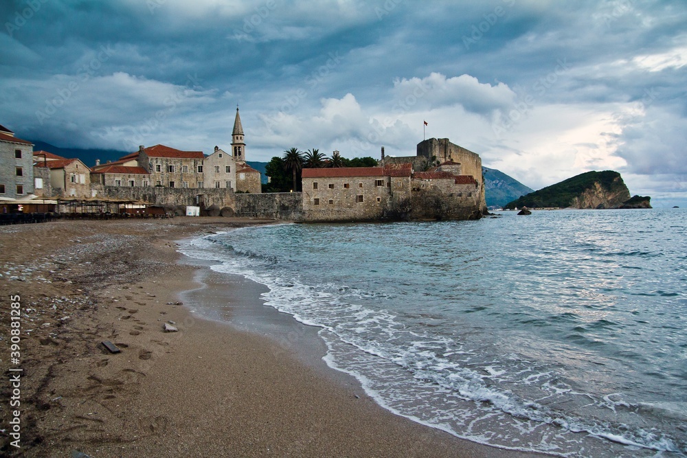 Grey houses with tile roofs and a cafe on the sandy beach of Adriatic Sea in the old town of Budva and against cloudy sky in Montenegro.