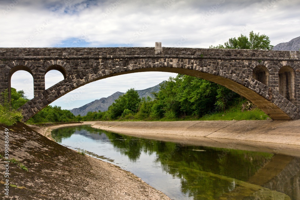 A stone bridge over the artificial channel with concrete embankment surrounded with green bushes and mountains in the background in Bosnia and Herzegovina.