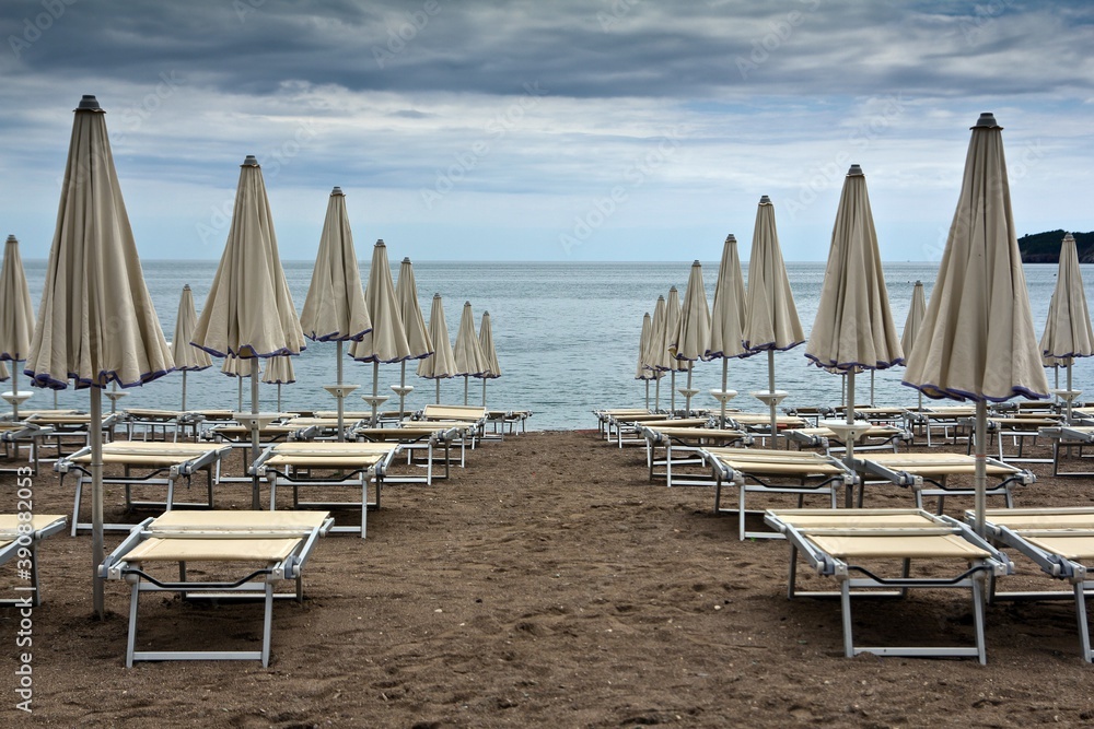 Many identical sunbeds with closed umbrellas on sandy beach of Adriatic sea against cloudy sky in Becici, Montenegro.