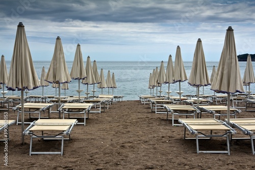 Many identical sunbeds with closed umbrellas on sandy beach of Adriatic sea against cloudy sky in Becici, Montenegro. © Алексей Мараховец